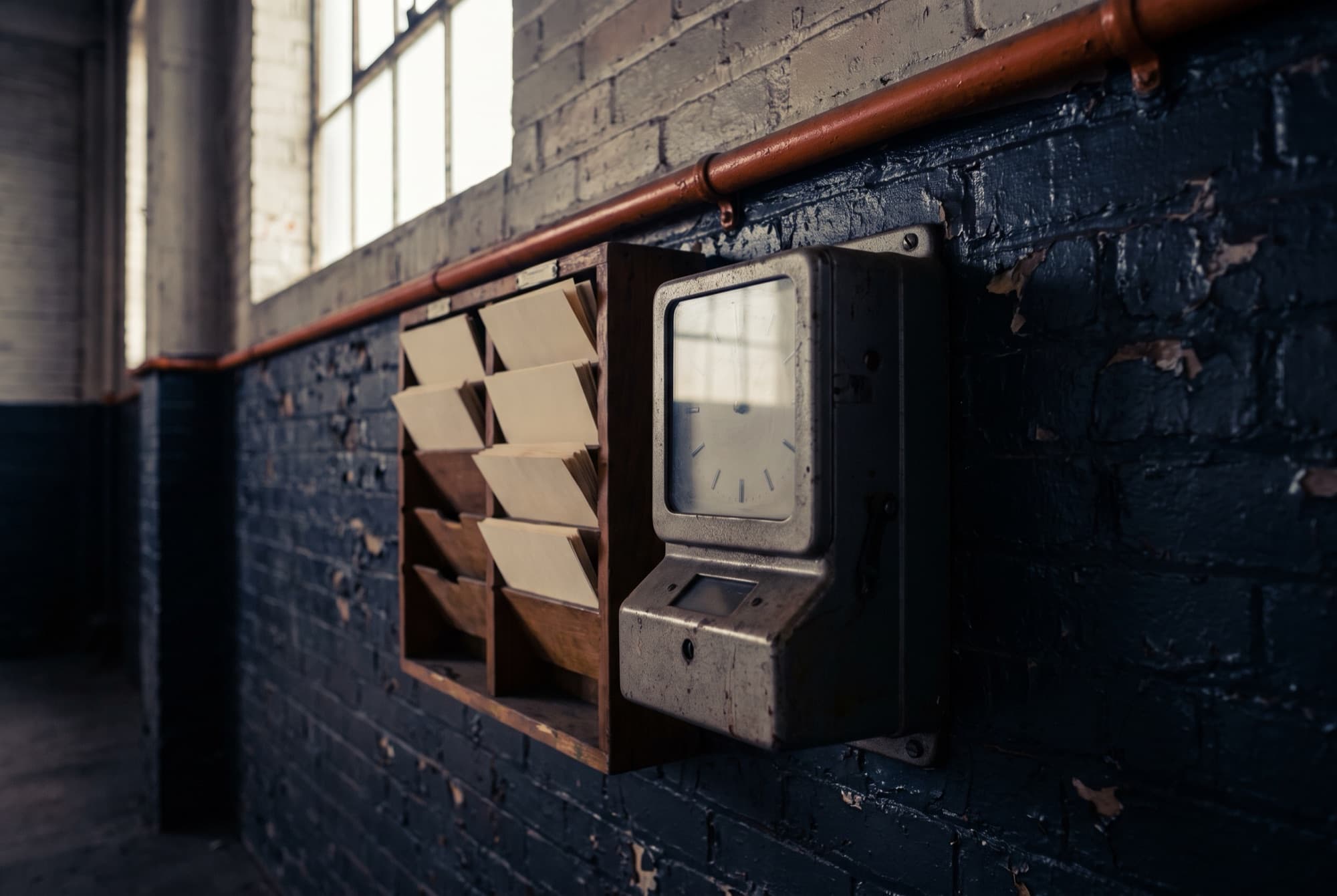A vintage punch time clock beside a rack of blank time cards on a dark brick wall in an empty factory corridor at dawn