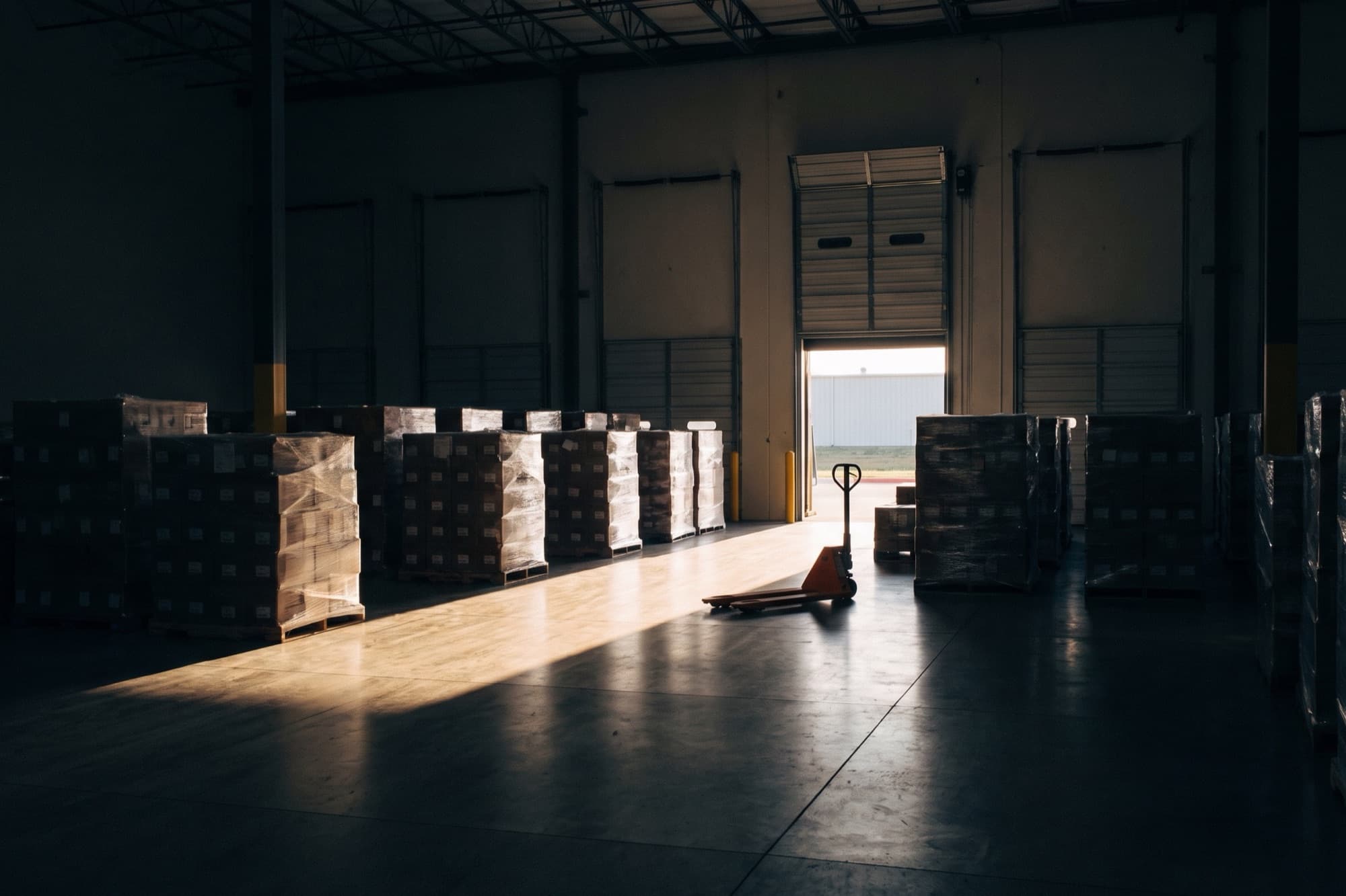 Shrink-wrapped pallets staged in a South Texas cross-dock warehouse, with morning sun falling through an open loading door