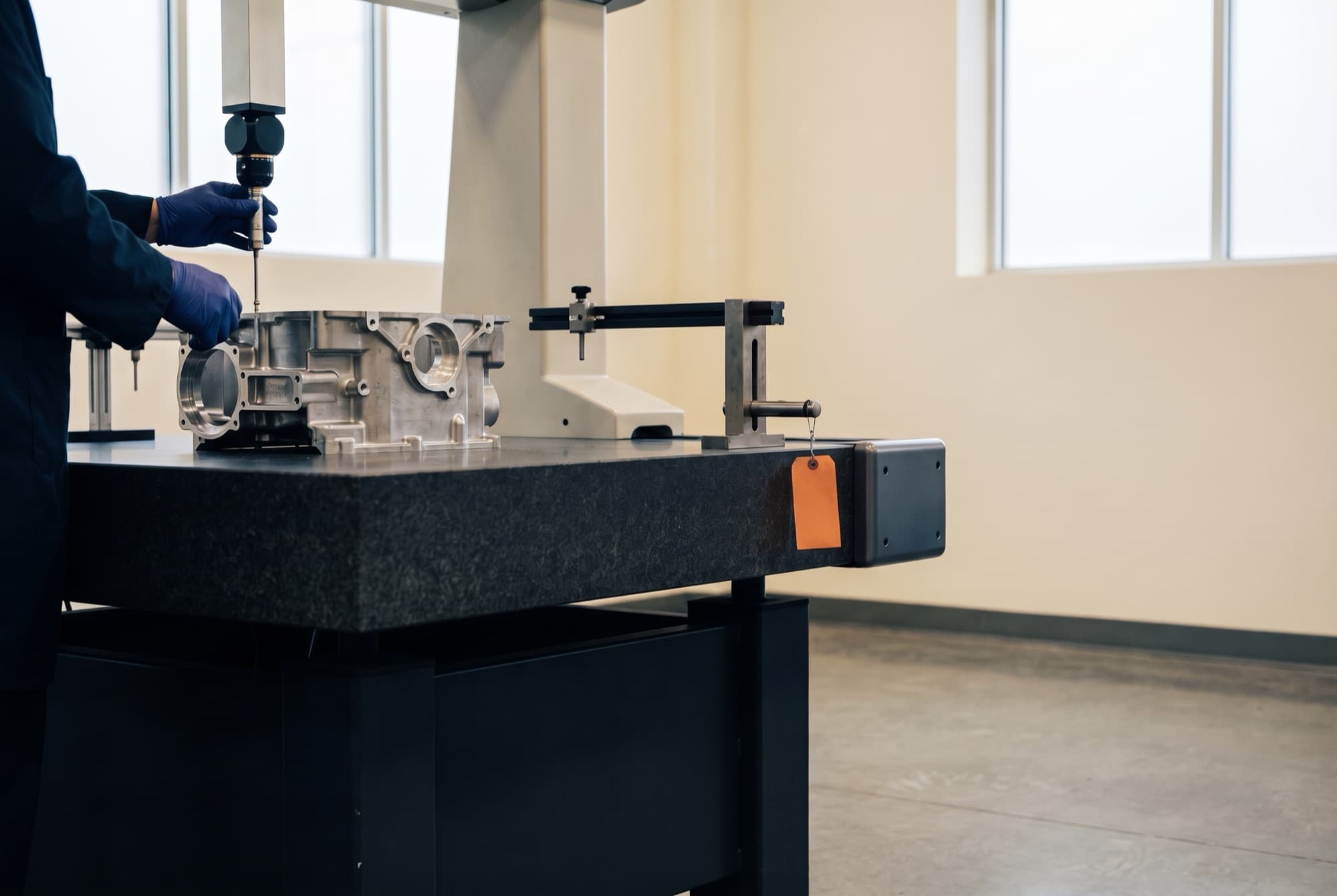 Gloved hands guiding the probe of a coordinate measuring machine over a machined aluminum housing in a factory quality lab