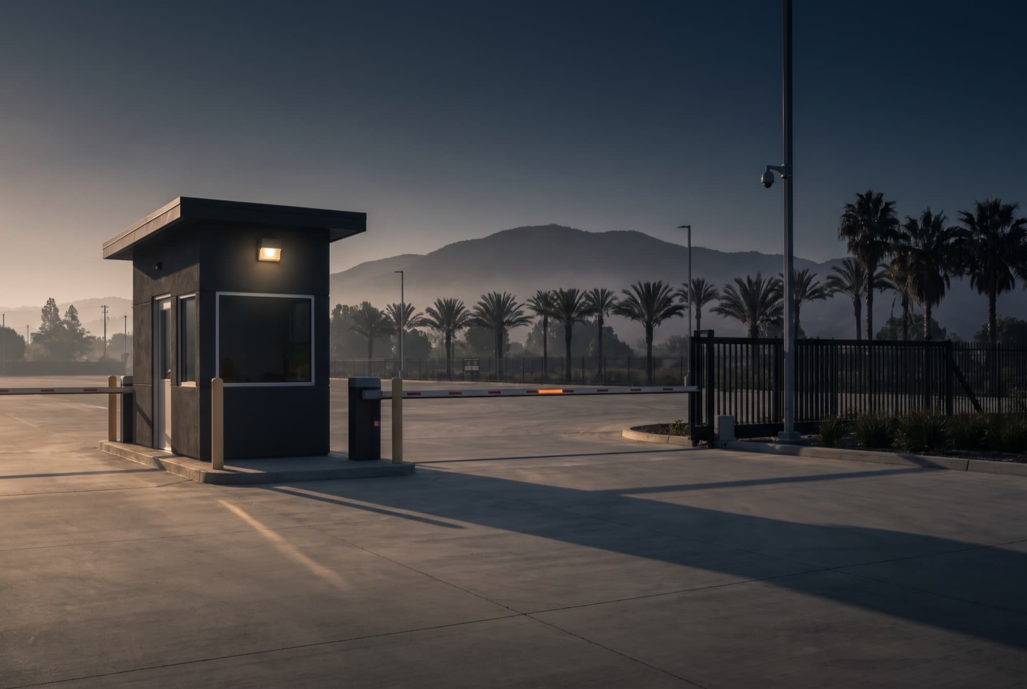A guardhouse with a lowered boom barrier at the entrance of an industrial park at dawn, palm trees and a mountain ridge behind