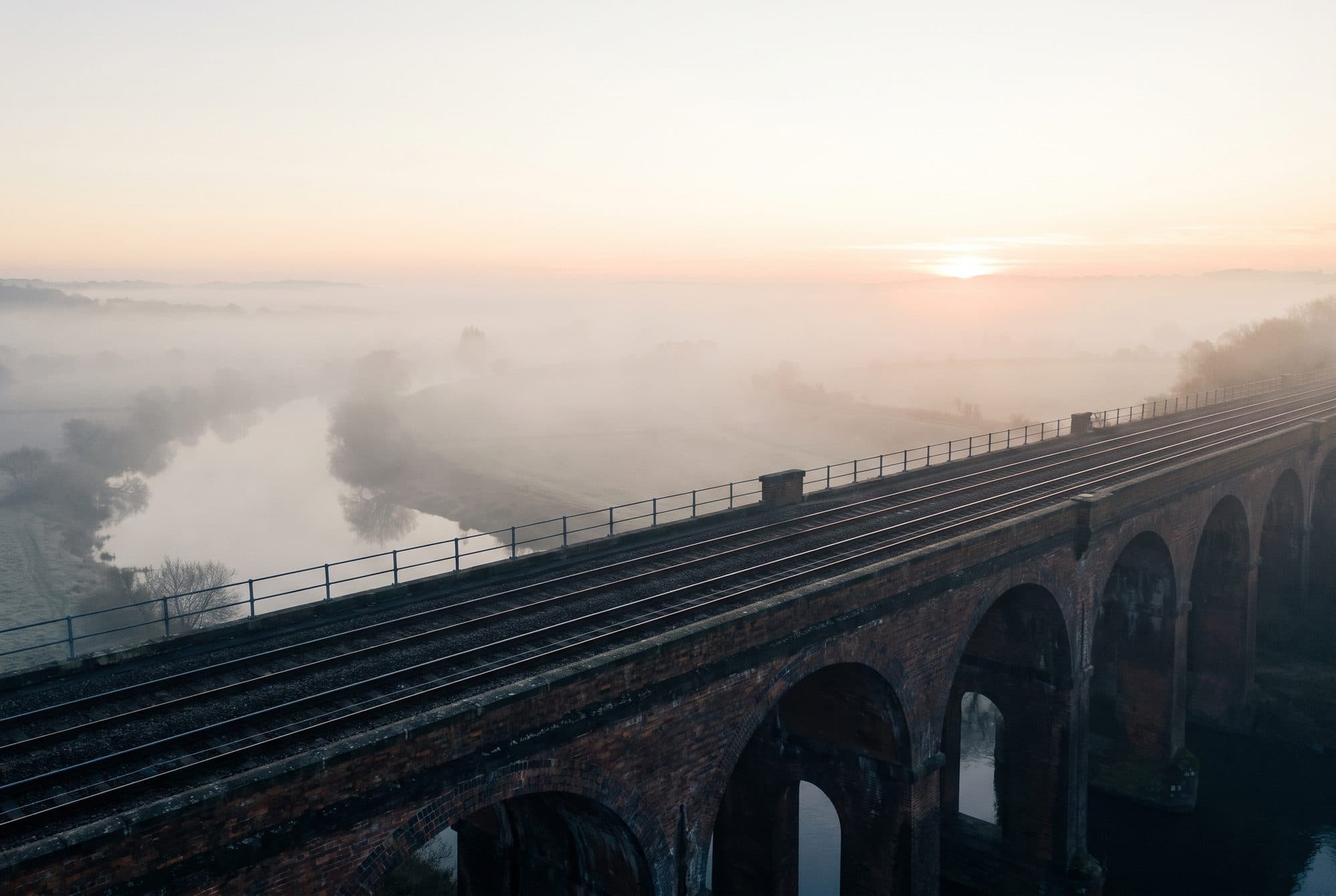 A Victorian brick railway viaduct over a misty river valley at dawn, empty tracks catching the first light